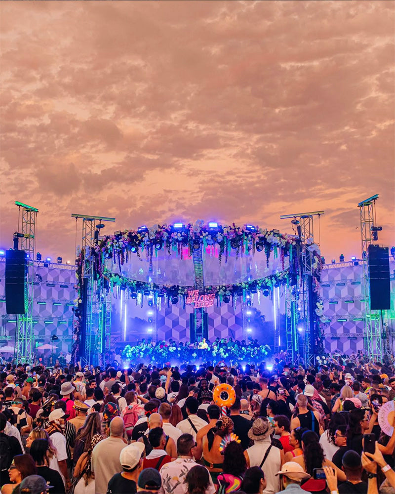 Crowd dancing under a circular stage decorated with flowers and lights at sunset during We Belong Here Festival.