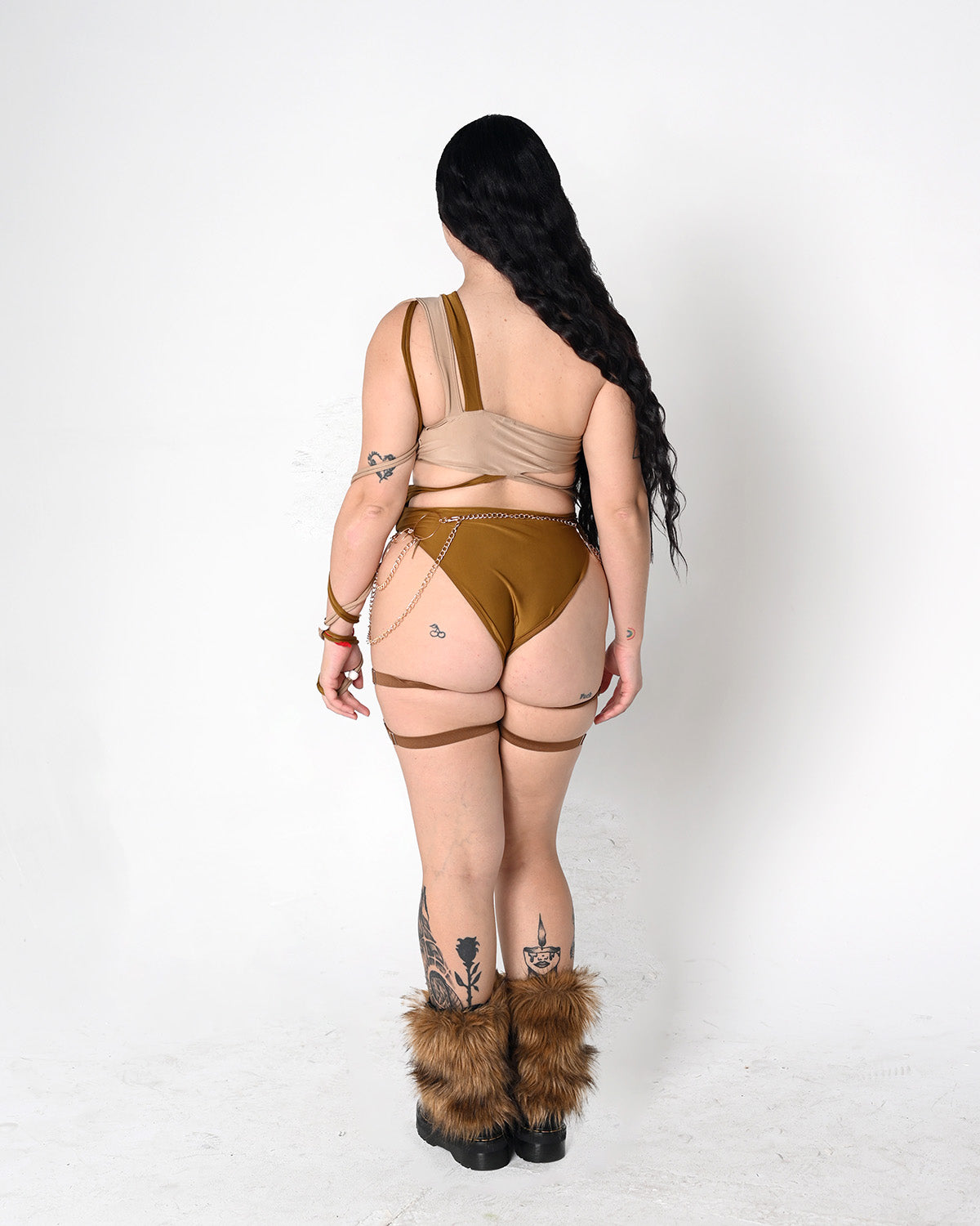 A woman is shown from behind wearing a beige and brown rave bodysuit set with garter straps, furry brown boot covers, and long black hair, standing against a plain white studio background.