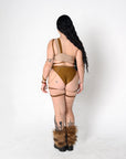 A woman is shown from behind wearing a beige and brown rave bodysuit set with garter straps, furry brown boot covers, and long black hair, standing against a plain white studio background.