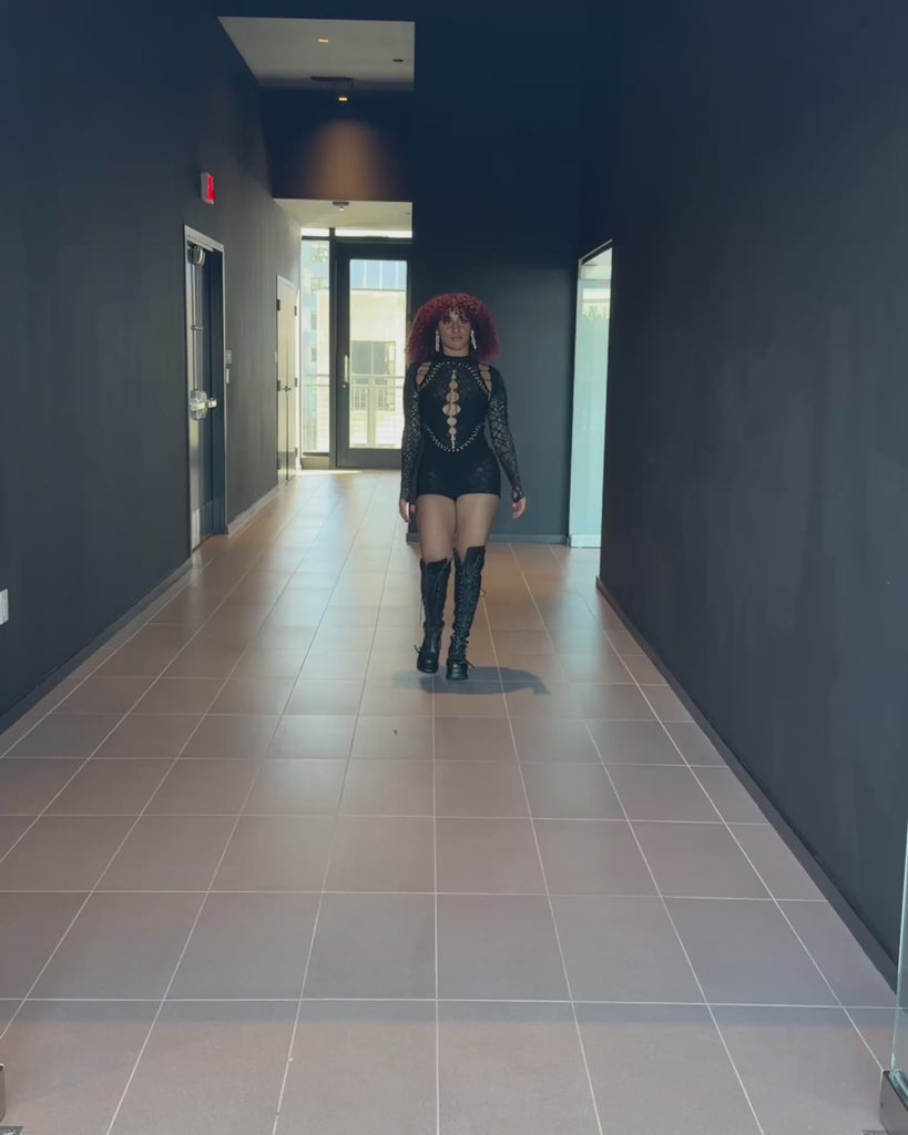 Model wearing a black mesh rave jumpsuit walking indoors, demonstrating fit through the hips and legs and how the fabric holds shape while moving.
